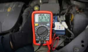 Ręka w czarnej rękawiczce trzyma multimetr pokazujący 12.72V. To pokazuje, jak sprawdzić napięcie 12V miernikiem przy akumulatorze samochodowym.