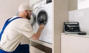 Serwisant sprawdza, czy nie ma urwanego bębna w pralce. Obok leży walizka z narzędziami.