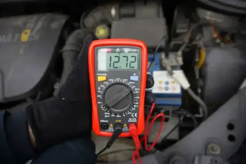 Ręka w czarnej rękawiczce trzyma multimetr pokazujący 12.72V. To pokazuje, jak sprawdzić napięcie 12V miernikiem przy akumulatorze samochodowym.