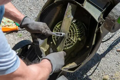 Ręce w rękawiczkach odkręcają nóż kosiarki, przygotowując ją do przeróbki na aerator.