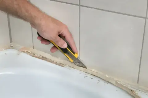 Dłoń z nożem odcina stary silikon przy wannie. Poznaj, jak usunąć stary silikon krok po kroku.