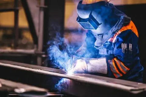 Spawacz w masce i rękawicach łączy metalowe elementy migomatem, tworząc snop iskier i niebieski dym. Dobór odpowiedniego gazu do migomatu jest kluczowy.