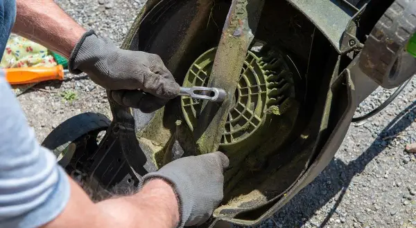 Ręce w rękawiczkach odkręcają nóż kosiarki, przygotowując ją do przeróbki na aerator.
