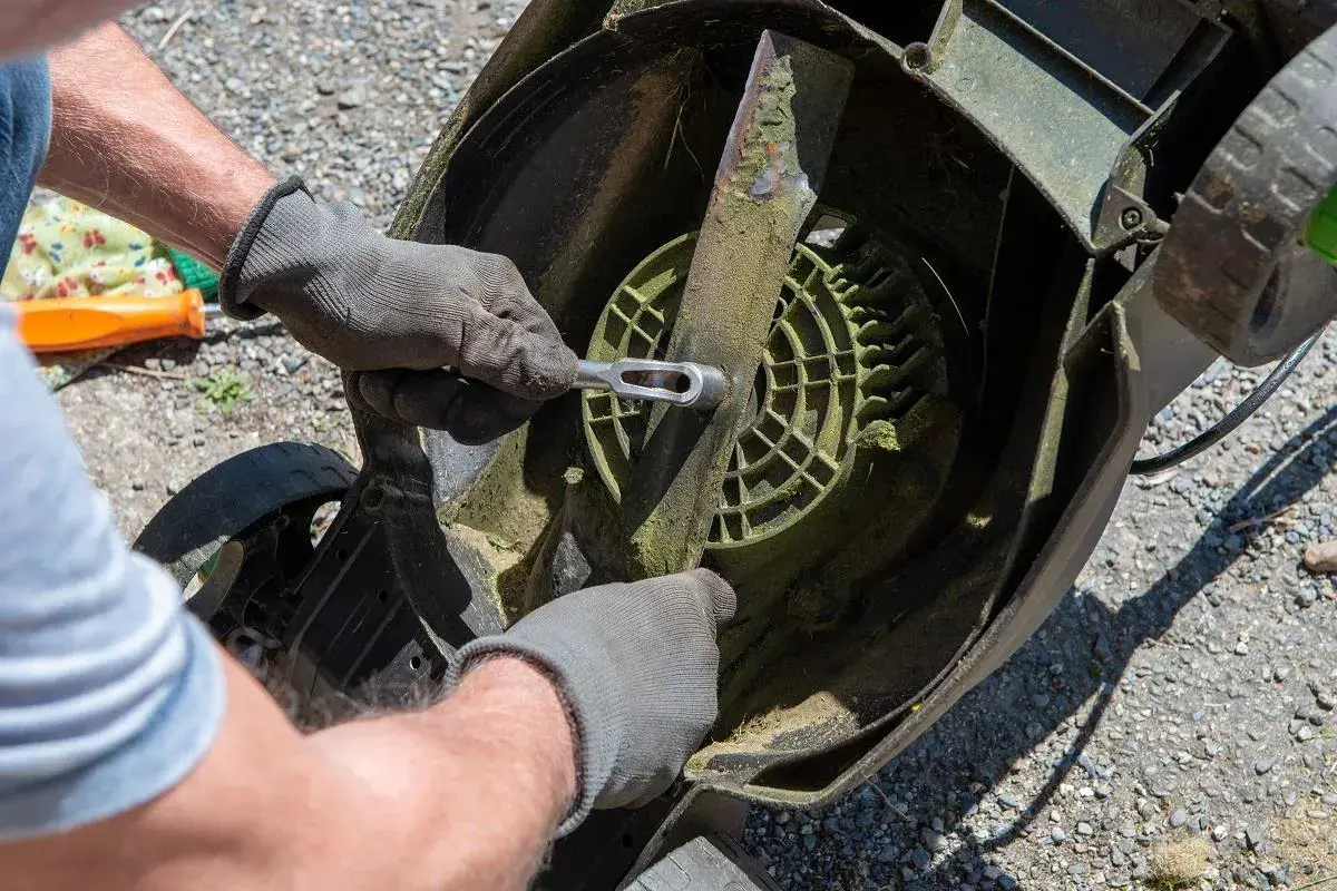 Ręce w rękawiczkach odkręcają nóż kosiarki, przygotowując ją do przeróbki na aerator.