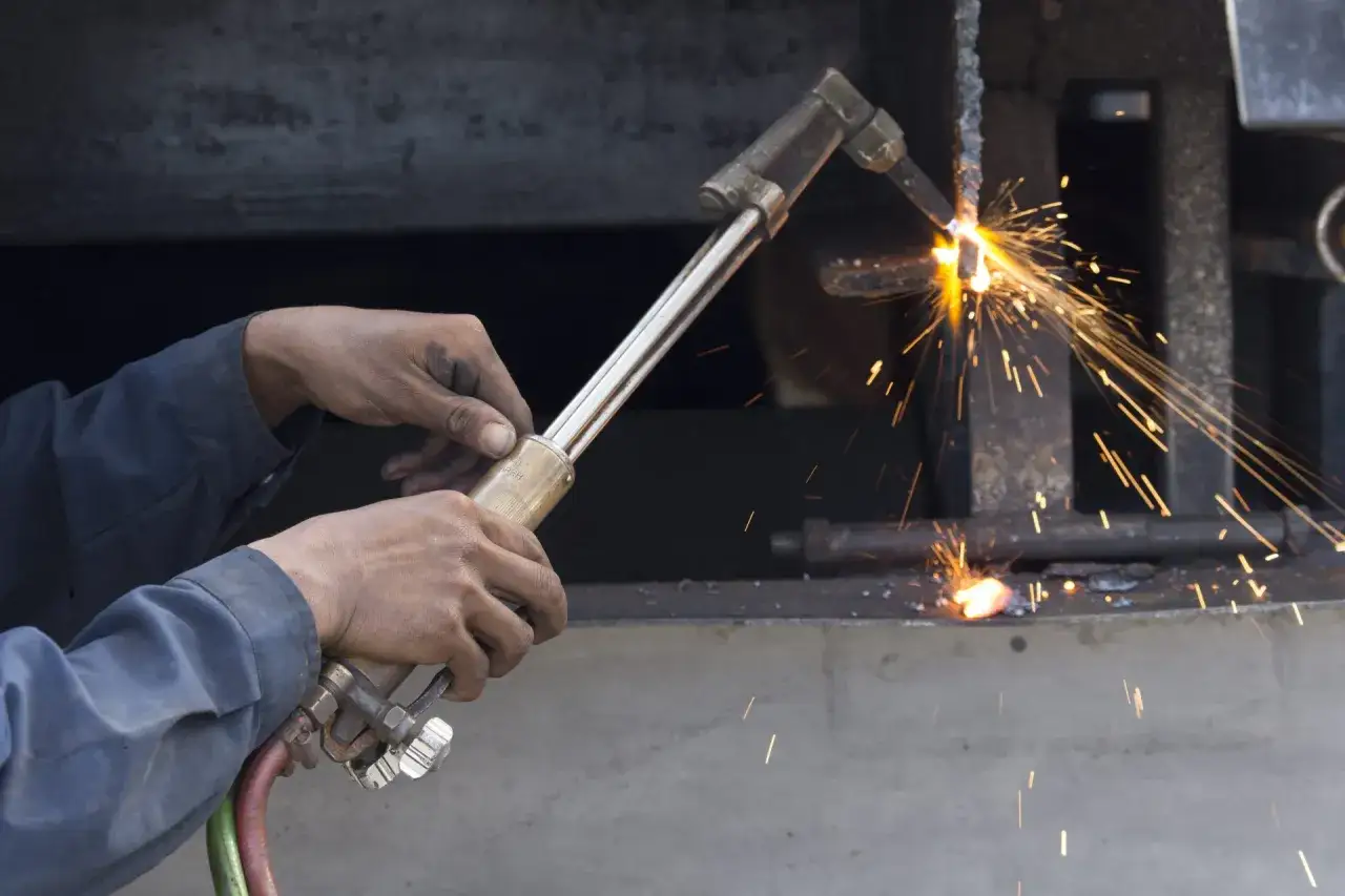 Ręce spawacza trzymają palnik, a iskry sypią się podczas spawania gazowego metalowych elementów.