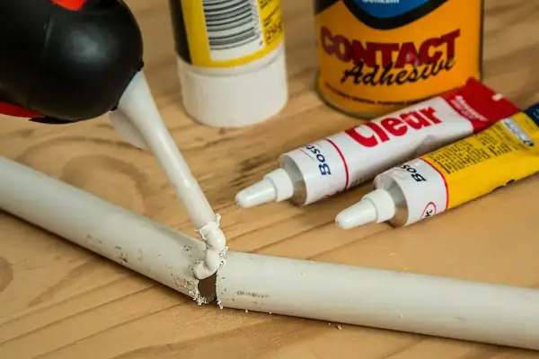 Aplikacja kleju z tubki na pękniętą plastikową rurkę. Obok leżą tubki z klejem i puszka "Contact Adhesive", pokazujące, czym skleić plastik.